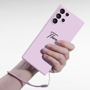 Puede incluir: Una funda de teléfono rosa claro con una inscripción negra que dice "Thom." La funda tiene una protuberancia de cámara con cinco lentes negros.