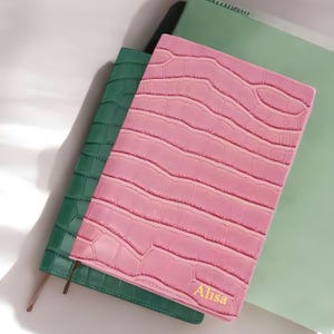 Könnte beinhalten: Zwei Notizbücher mit Krokodilprägung. Eines ist rosa und das andere grün. Das rosa Notizbuch hat den Namen "Alisa" auf dem Einband geprägt.