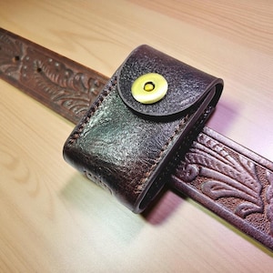 May include: A dark brown leather belt case with a gold-colored snap closure, attached to a brown leather belt with embossed floral designs. The case is stitched with a contrasting thread.