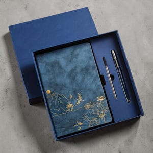 May include: A blue gift set containing a notebook and two pens. The notebook has a textured cover with a gold embossed landscape design. The set is presented in a matching blue box, ideal for gifting.