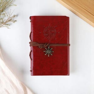 Puede incluir: Un diario de cuero rojo con un diseño de rosa de los vientos en relieve en la portada. El diario está encuadernado con un cordón marrón y presenta un dije decorativo de timón. El diario está cerrado.