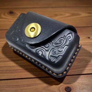 May include: Black leather case with an embossed floral design and a brass button closure. The rectangular case has a flap secured by a circular brass button. The edges are stitched with light brown thread.