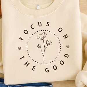 May include: Cream-colored sweatshirt with the text "FOCUS ON THE GOOD" in black, surrounding a floral design. The sweatshirt is paired with blue denim jeans and a brown hat, creating a casual, everyday look.