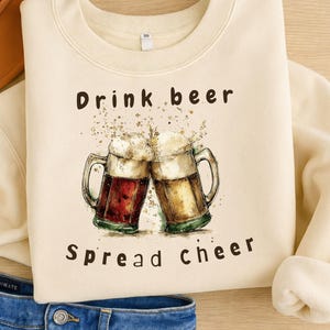 May include: Cream-colored sweatshirt with the text "Drink beer Spread cheer" above a watercolor illustration of two beer mugs clinking together. The mugs are filled with beer and foam. The sweatshirt is paired with blue jeans and a brown hat.