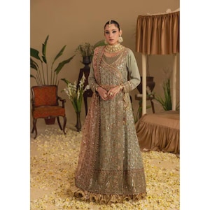 May include: A woman wearing a sage green and gold embroidered gown with long sleeves and a matching sheer shawl. The gown features intricate floral patterns and gold embellishments. She is wearing gold jewelry, including a necklace, earrings, and rings. The background includes a vintage chair and floral arrangements.