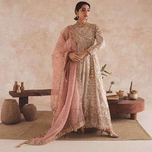 May include: A formal, floor-length gown in a light beige colour, adorned with intricate gold embroidery. The dress features long sleeves and a matching pink dupatta. A gold necklace and earrings complete the ensemble. Suitable for special occasions.