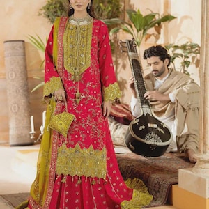 Könnte beinhalten: Eine Frau in einem rot-goldenen bestickten Kleid mit passender Handtasche und Schal. Das Kleid hat aufwendige goldene Details und einen gestuften Rock. Im Hintergrund spielt ein Mann Sitar.