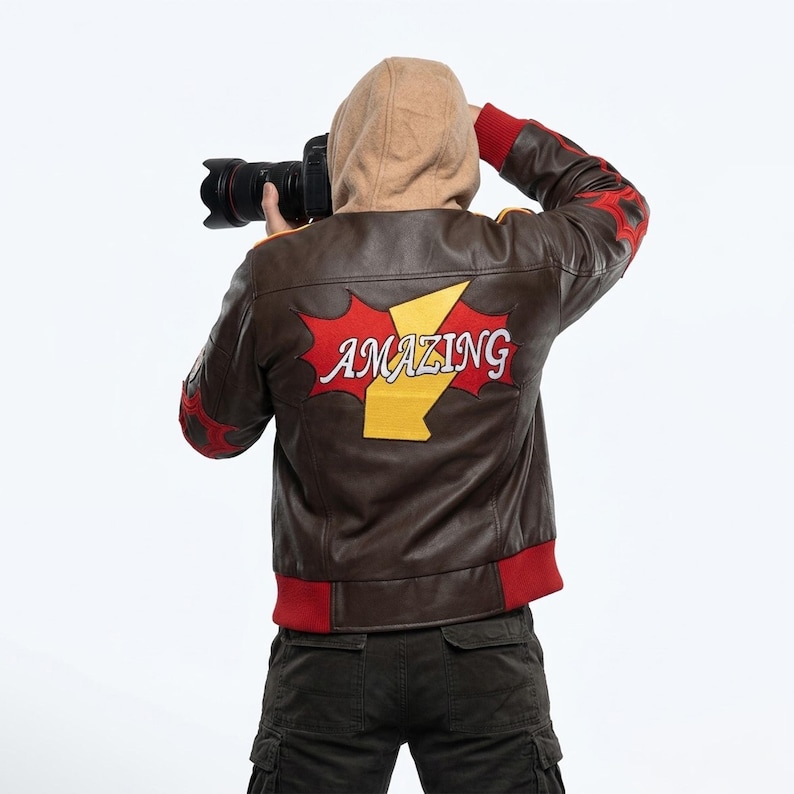 May include: A brown leather jacket with a tan hood, red ribbed cuffs, and a red hem. The back features a large red starburst patch with a yellow lightning bolt and the word AMAZING in white letters. The sleeves have red starburst accents.