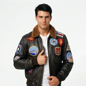 May include: A brown leather bomber jacket with multiple patches, a brown fur collar, and brown ribbed cuffs. The jacket has a zipper closure and a white t-shirt underneath. Patches include the word "Dallas" and the word "Tomcat".