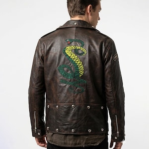Può includere: Giacca in pelle marrone con un grande disegno di serpente ricamato sul retro. La giacca ha borchie color argento, chiusura con zip e una piccola toppa sulla manica. Il serpente è verde e giallo.