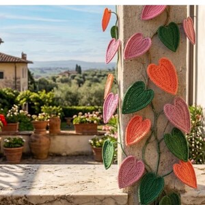 Puede incluir: Una vid decorativa de ganchillo con hojas en forma de corazón en tonos rosa, naranja y verde. La vid está unida a una columna de piedra, con un fondo exterior escénico con plantas en macetas y un paisaje distante.