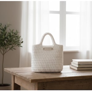 May include: A white crochet tote bag with two handles, sitting on a wooden table. The bag has a rectangular shape and a small tag with text. The background includes a window and a small plant.
