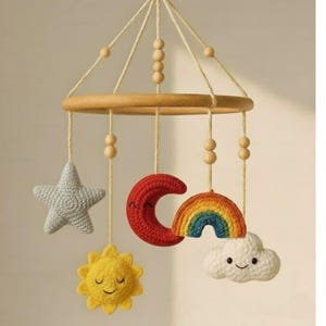 Può includere: Mobile per bambini fatto a mano con un anello di legno e forme all'uncinetto sospese. Le forme includono una stella grigia, una luna crescente rossa, un sole giallo, un arcobaleno e una nuvola bianca con un volto sorridente. Il mobile è sospeso con spago color naturale.