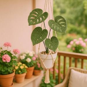 Puede incluir: Un macetero colgante de macramé con una maceta de cerámica blanca que contiene una planta de Monstera verde tejida a ganchillo. La planta tiene hojas grandes y detalladas. El fondo incluye otras plantas en macetas y una barandilla de madera.