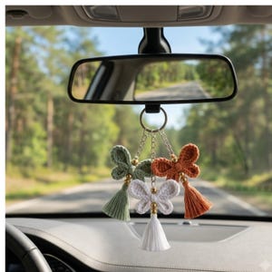 May include: A car rearview mirror hanging ornament featuring three crocheted butterfly shapes in sage green, white, and burnt orange. Each butterfly is accented with a small gold-toned bead and a matching colored thread tassel hanging from a silver chain.