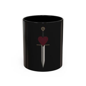 Puede incluir: Taza de cerámica negra con un corazón burdeos atravesado por una espada plateada. La empuñadura de la espada es dorada. La taza es cilíndrica y tiene un acabado brillante. El diseño está centrado en la taza.