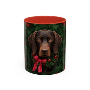 May include: A ceramic coffee mug featuring a brown dog with a Christmas wreath and red bow. The interior of the mug is red, and the background is a dark green with red ribbons. A festive holiday design.