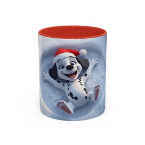 Puede incluir: Taza de cerámica blanca con interior rojo. La taza presenta un cachorro dálmata de dibujos animados con un gorro de Papá Noel, tumbado en una escena nevada. El cachorro tiene manchas negras y sonríe con los brazos extendidos.