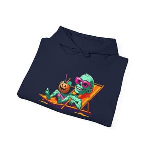 Puede incluir: Una sudadera con capucha azul marino con una ilustración de dibujos animados de un zombi verde con gafas de sol rosas, relajándose en una tumbona con un cóctel de calabaza. El diseño tiene colores brillantes y un tema de Halloween.