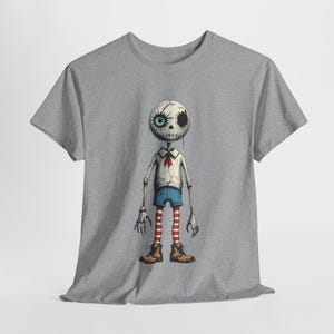 Puede incluir: Una camiseta gris con una ilustración de dibujos animados de una figura esquelética. La figura tiene una cabeza cosida con un ojo azul y un ojo negro, que lleva una camisa blanca, pantalones cortos azules y calcetines a rayas rojas y blancas. La figura lleva botas marrones.