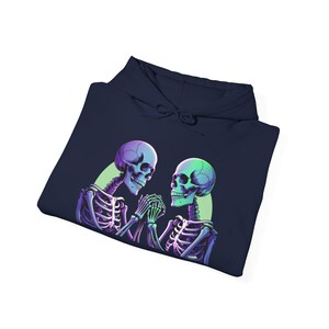 Puede incluir: Sudadera con capucha azul marino con un gráfico de dos esqueletos tomados de la mano. Los esqueletos tienen acentos morados y verdes, con un brillo verde a juego alrededor de sus cuellos. El diseño está centrado en la parte delantera de la sudadera.