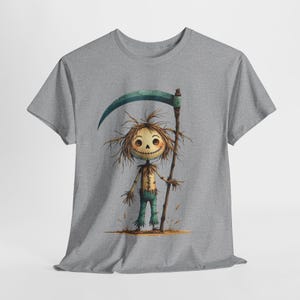 Puede incluir: Camiseta gris con una ilustración caprichosa de un espantapájaros. El espantapájaros tiene una cara cosida, pelo de paja y sostiene una guadaña. Viste pantalones azules y una camisa cosida. El diseño está sobre un fondo neutro.