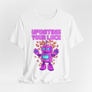 Puede incluir: Camiseta blanca con un robot de dibujos animados morado y el texto "UPDATING YOUR LUCK" en rosa. El robot está rodeado de monedas de oro y tiene una pantalla que dice "YOUR LUCK".
