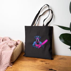 Può includere: Borsa tote nera con una grafica di un cane con una maschera e una motosega rosa. Il cane indossa una camicia blu e un grembiule rosa. La borsa ha manici neri ed è realizzata in materiale resistente.