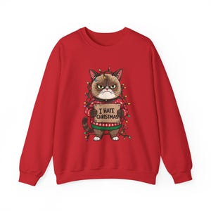 Puede incluir: Sudadera roja con un gato gruñón que lleva un jersey navideño y luces de colores. El gato sostiene un cartel que dice "I HATE CHRISTMAS". El gato es marrón y blanco con una expresión de mal humor. El jersey tiene un estampado rojo y verde.