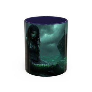 May include: A white ceramic mug with a dark blue interior. The exterior features a fantasy illustration of a mermaid with green eyes and scales, sitting near a shipwreck under a stormy sky. The mug is designed for hot beverages.