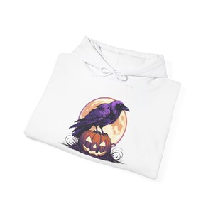 Puede incluir: Sudadera con capucha blanca con un diseño de Halloween. El gráfico muestra un cuervo morado sobre una calabaza naranja tallada, con una luna llena de fondo. La sudadera está hecha de un material suave.