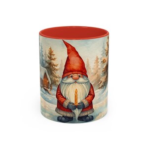 May include: A ceramic mug with a red interior and a winter scene design. The design features a gnome wearing a red hat and coat, holding a candle. The background shows a snowy landscape with a cabin and pine trees.