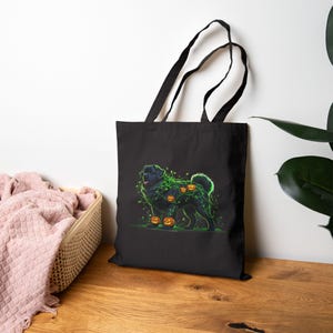 May include: Black tote bag featuring a Halloween-themed illustration. The design depicts a black dog with green vines and orange jack-o'-lanterns. The bag has black handles and is made of a durable fabric, suitable for carrying items.