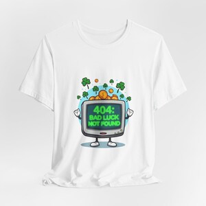 Puede incluir: Camiseta blanca con un televisor de dibujos animados que muestra "404: BAD LUCK NOT FOUND" en verde. El televisor tiene brazos y piernas, con monedas de oro y tréboles encima. Un diseño gráfico divertido.