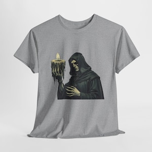 Puede incluir: Una camiseta gris con un gráfico de una figura con túnica sosteniendo una vela encendida. La figura está representada en tonos negros y grises, con la vela emitiendo una llama amarilla. El diseño tiene un tema gótico o de fantasía.
