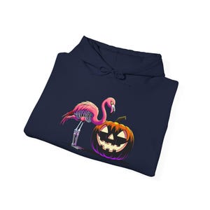 Puede incluir: Una sudadera con capucha azul marino con un diseño de Halloween. El diseño incluye un flamenco rosa con un esqueleto, junto a una calabaza de Halloween. La calabaza tiene una cara tallada con un contorno púrpura. El flamenco tiene un cuerpo rosa y un esqueleto.