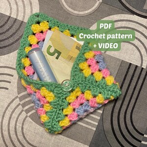 Peut inclure: Une pochette en crochet avec un motif de couleur vert, rose, jaune et bleu. La pochette est ouverte et contient un petit tube bleu et de l'argent liquide. Le texte "PDF Crochet pattern + VIDEO" est sur un fond blanc.