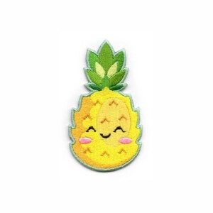 Puede incluir: Un parche bordado con forma de piña con cuerpo amarillo y hojas verdes. La piña presenta una cara sonriente con los ojos cerrados, una pequeña boca curva y dos mejillas circulares de color rosa, todo bordeado con un hilo azul claro.