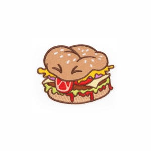 Puede incluir: Un parche bordado con forma de hamburguesa que presenta un pan color tostado con semillas de sésamo blancas, una carne marrón, lechuga verde, tomate rojo, queso amarillo y una rodaja de cebolla morada, todo delineado con hilo marrón oscuro.