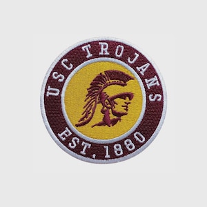 May include: A circular embroidered patch featuring a yellow center with a maroon Trojan helmet profile. The maroon border contains the white text USC TROJANS and EST. 1880, all outlined in a thin white stitched edge on a plain background.
