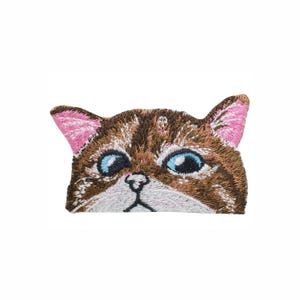 Puede incluir: Un parche bordado con forma de cabeza de gato con pelaje marrón y blanco, ojos azul brillante, nariz negra y orejas rosadas por dentro. El diseño presenta costuras detalladas que capturan los rasgos faciales de un gato atigrado.