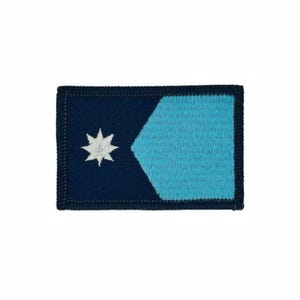 May include: A rectangular embroidered patch featuring a dark navy blue background on the left side with a white eight-pointed star. The right side consists of a light blue pentagonal shape with a dark navy blue border surrounding the entire patch.