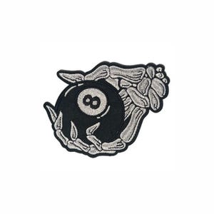May include: An embroidered patch featuring a skeletal hand gripping a black billiard ball with a white circular center containing the number eight. The design uses black and white thread to create a high-contrast graphic of a bony hand holding the ball.