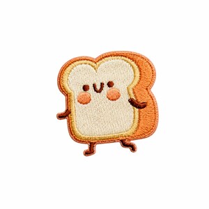 May include: An embroidered patch shaped like a slice of toasted bread with a smiling face. The centre is cream-coloured with two pink cheeks, while the crust is a golden brown. The patch features small brown arms and legs in a walking pose.