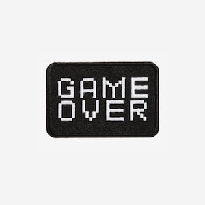 May include: A rectangular embroidered patch with a black background and white pixelated text that reads GAME OVER. The patch features a clean, stitched border and a blocky, retro-inspired font style, designed for application onto fabric surfaces.