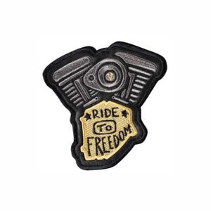 May include: An embroidered patch featuring a V-twin motorcycle engine design in shades of black, grey, and silver. A central gold-colored shield shape contains the black embroidered text RIDE TO FREEDOM with two small black stars on either side.
