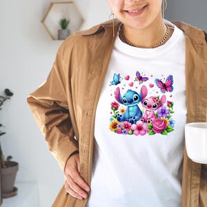 May include: White t-shirt featuring a colorful graphic of two cartoon characters surrounded by flowers, butterflies, and hearts. The characters are blue and pink, with large ears. The design is vibrant and detailed.