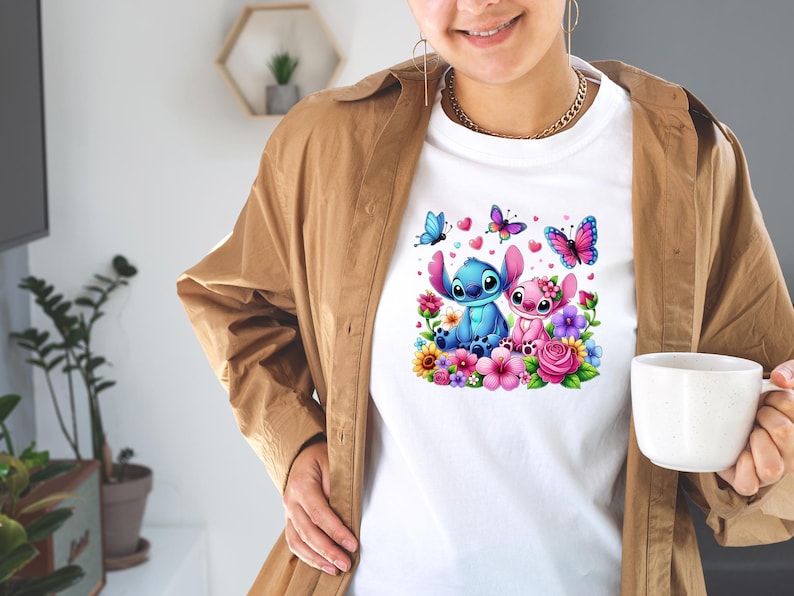 May include: White t-shirt featuring a colorful graphic of two cartoon characters surrounded by flowers, butterflies, and hearts. The characters are blue and pink, with large ears. The design is vibrant and detailed.
