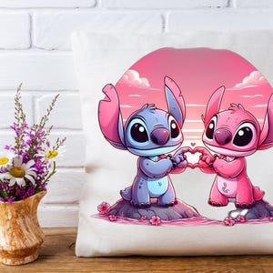 May include: A white pillow featuring a cartoon illustration of two alien-like characters holding a heart shape. One character is blue, the other pink, set against a pink and orange sunset background. The pillow is on a white surface.