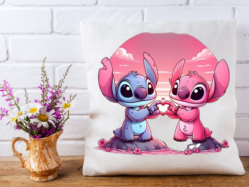 May include: A white pillow featuring a cartoon illustration of two alien-like characters holding a heart shape. One character is blue, the other pink, set against a pink and orange sunset background. The pillow is on a white surface.
